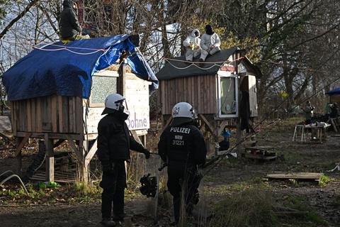 Polizisten stehen bei der Räumung von Lützerath mit Schilden vor Bretterhütten auf deren Dächern Demonstranten sitzen.