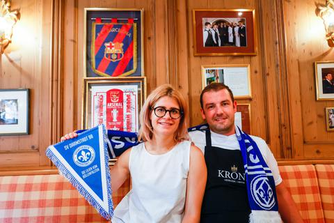 Hotelinhaberin Erika Kuntz und ihr Mann, Chef de Cuisine Fabio Daneluzzi, vor den Fotos von prominenten Persönlichkeiten und Fußballvereinen, die bereits Gäste waren. Neben den Wimpeln des FC Barcelona und Arsenal London soll nun auch der SV Darmstadt 98 ein Plätzchen finden. 